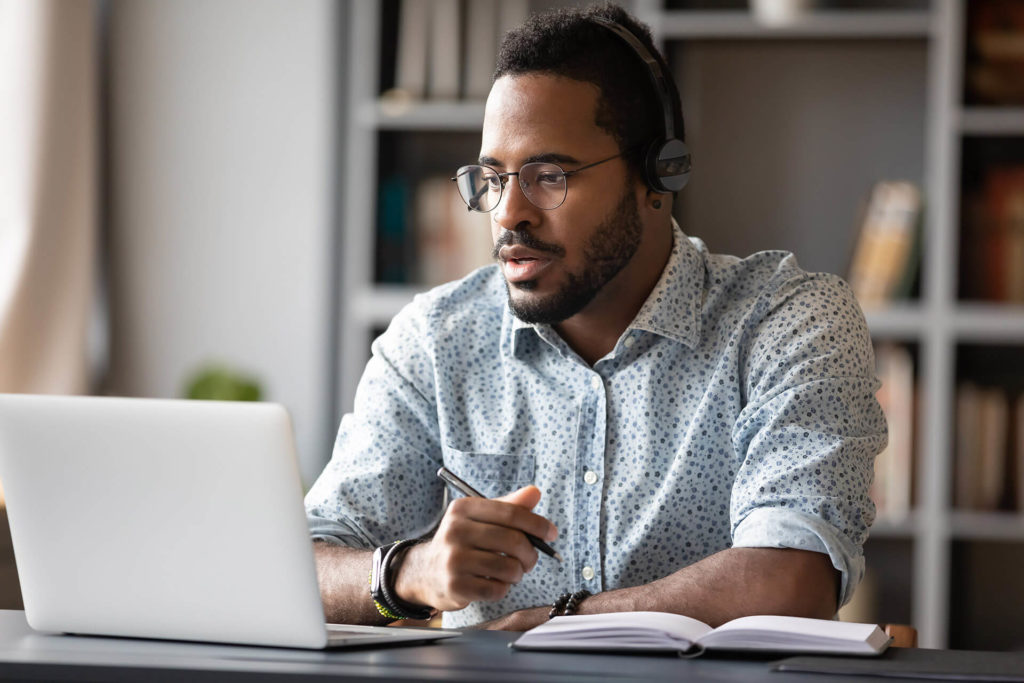 A concentrated male travel agent sits at his desk writing in a notebook and speaking through his headset to book the Sky Bird Travel & Tours travel deals.