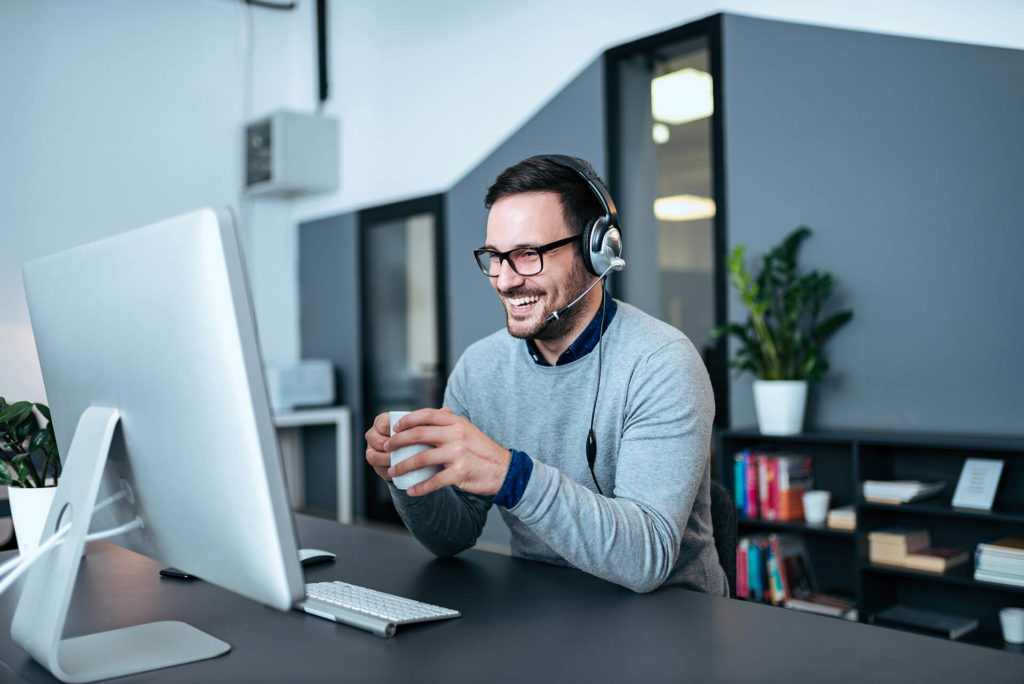 A smiling male travel agent holds a cup of coffee while talking into a headset connected to his computer, where he researches and books flights, hotels, activities, tours, cruises, and travel insurance for his clients through Sky Bird Travel & Tours.