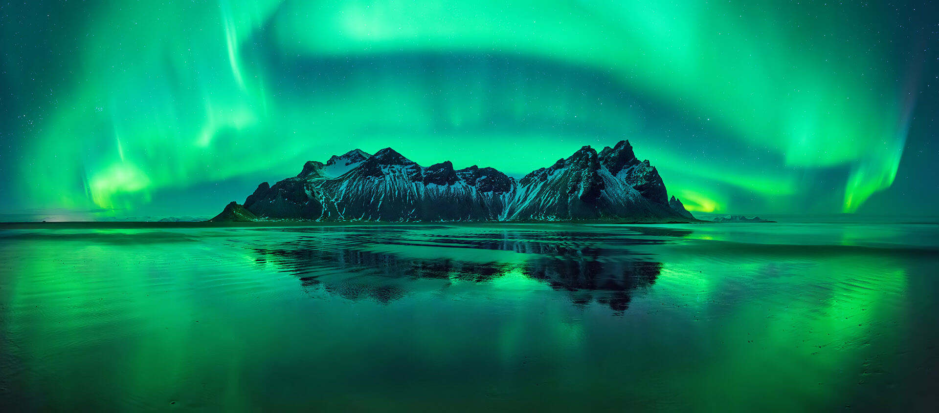 A tall and snowy mountain in Iceland surrounded by a beautiful ocean being cruised by Sky Bird Travel & Tours Sky Vacations. In the background, the beautiful natural phenomenon of the aurora borealis, or the northern lights, brightens the night sky with green hues.