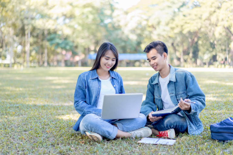 Two young international students sit on the grass in a park studying from a laptop and textbook comparing notes. They are on a cheap study abroad trip organized with the collaboration of the university and Sky Bird Travel & Tours.