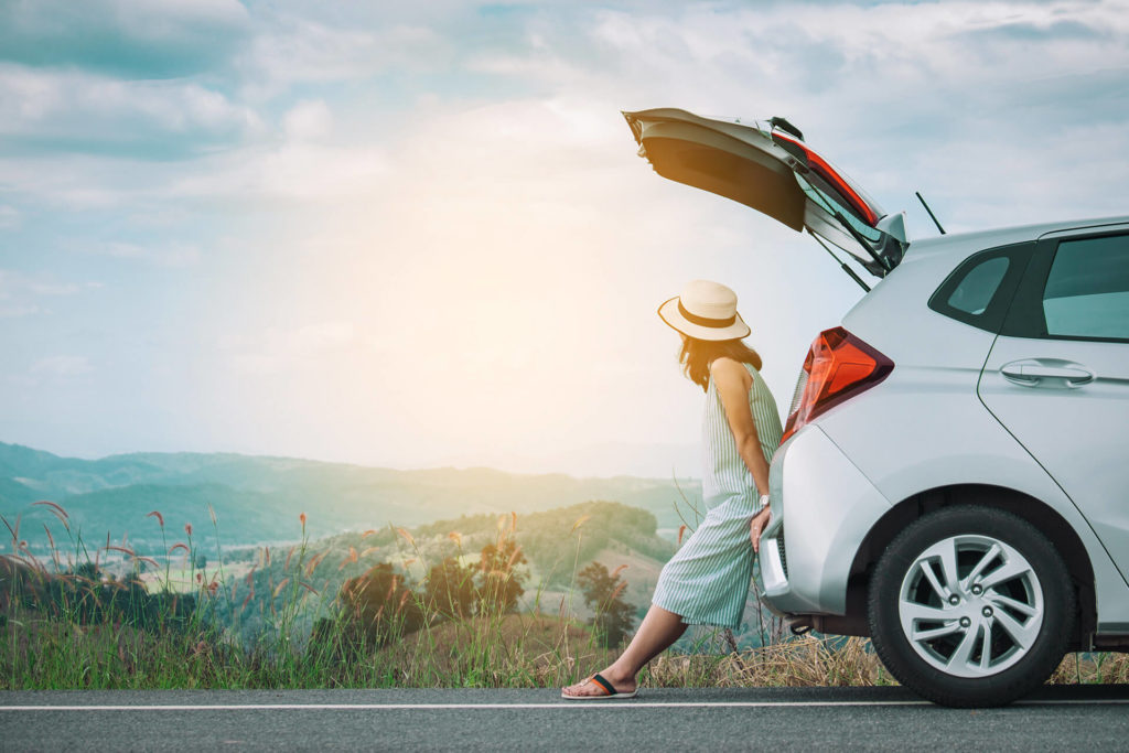 A woman traveler wearing a straw hat and sundress sits in the trunk of her rental car watching the peaceful sunrise over the hills and mountains, which she rented from Sky Bird Travel & Tours.