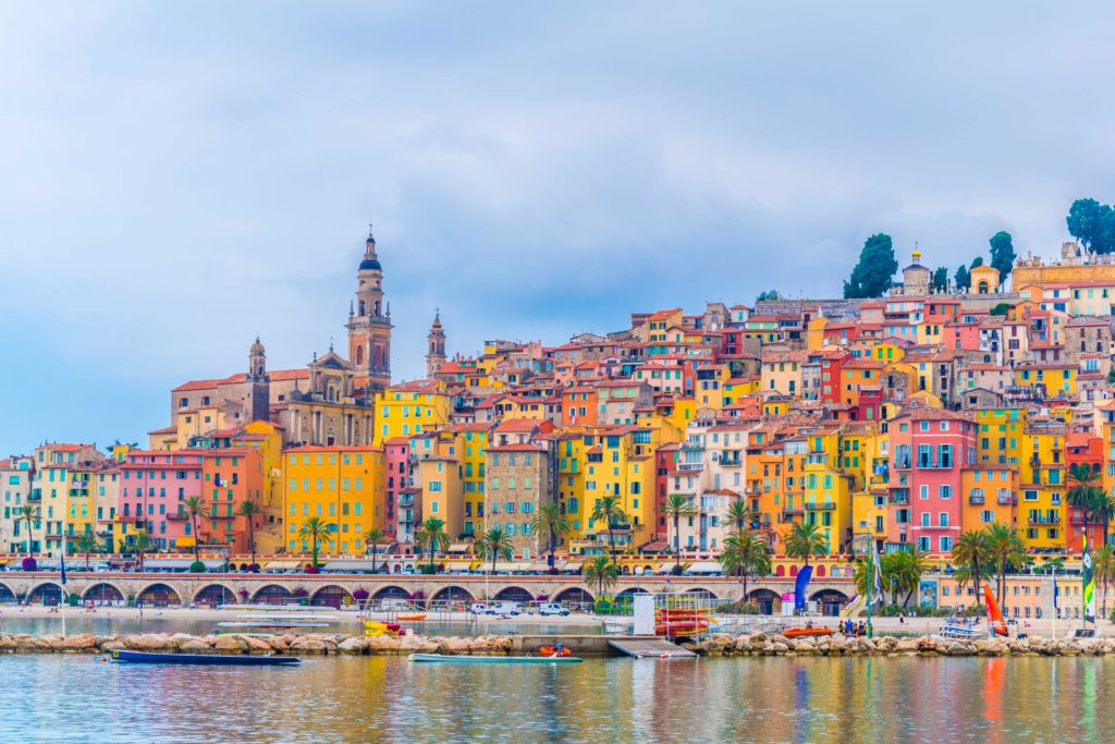 A travel picture of the beautiful blue sky and bright small townhouses in Menton, France. Sky Bird Travel & Tours recommends taking a summer vacation here in 2023.