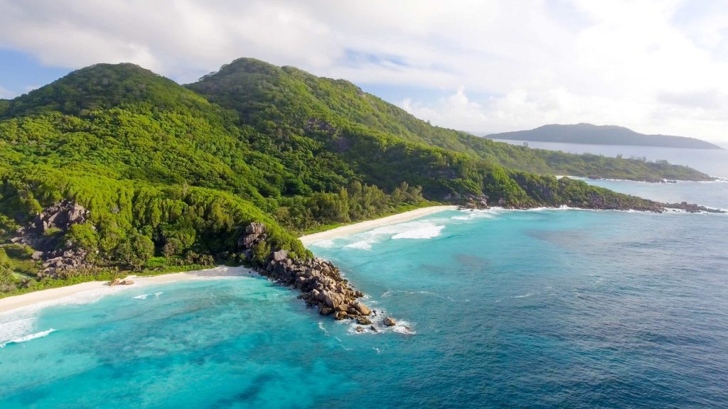 A beautiful wide shot of the lush green tropical La Digue Island of Seychelles in Africa. The waves of the blue ocean wash onto the sandy beach, the perfect summer destination.