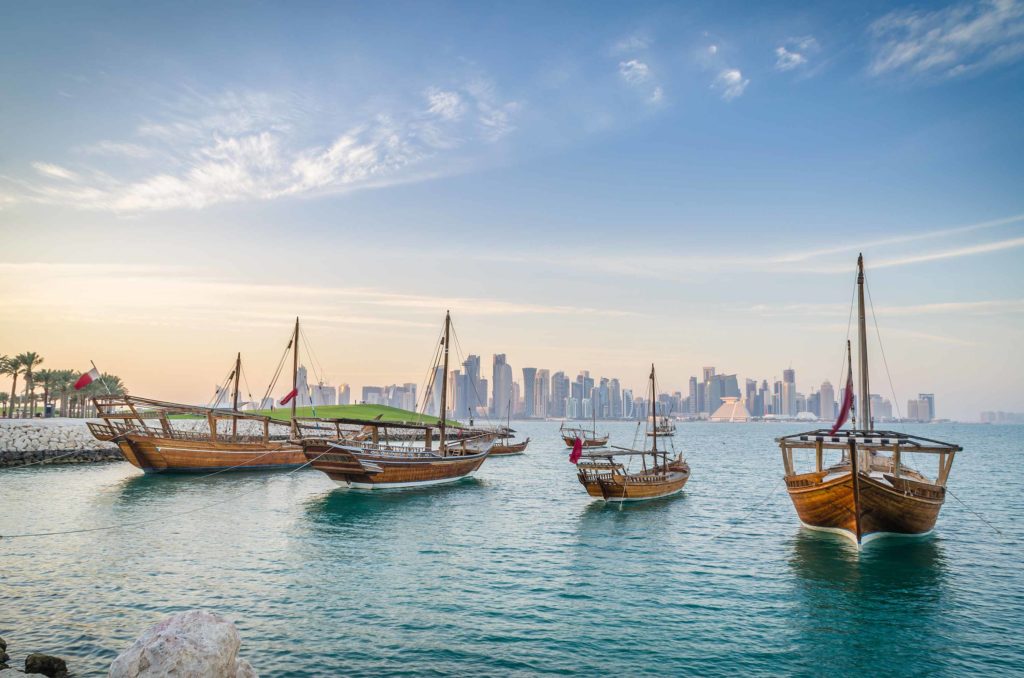 Four wooden boats with their sails lowered sit anchored in a blue bay, and there is an urban cityscape in the background across the water. Sky Bird Travel & Tours encourages their travel agents and travel advisors to book boat tours for clients