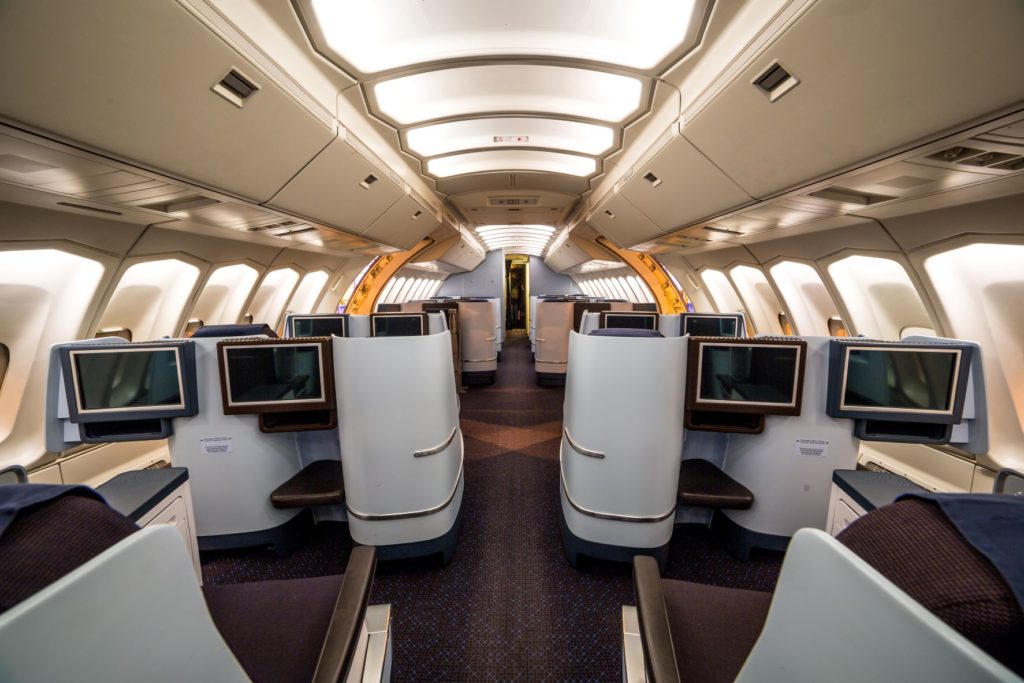 The interior of a luxury first-class cabin aboard an aircraft. Everything is white, and there is a lot of room between the aisles and seats. There are also televisions in front of every seat, a promise from the airline consolidator.