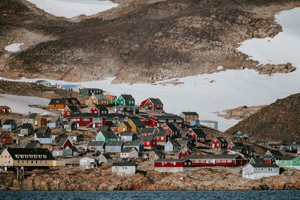 While the upper part of the photo is a bare rocky mountain and snow, the bottom half is a small village of colorful farmhouses. This is in an isolated town called Ittoqqortoormiit in Greenland and a top summer vacation destination by sky bird travel & tours.