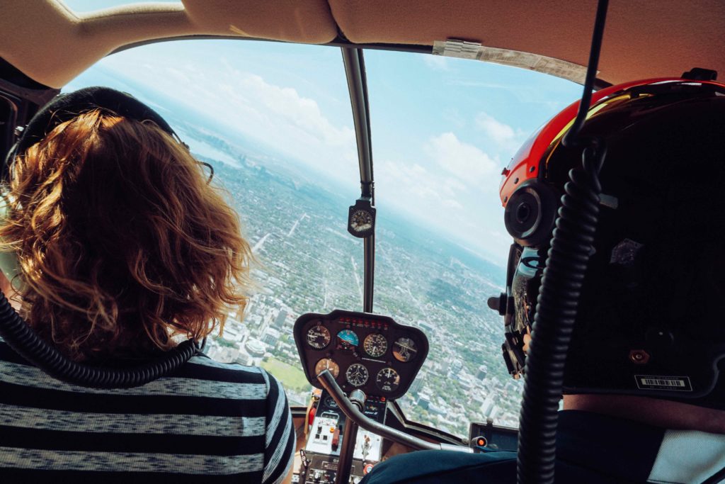 A woman with short hair sits in the drivers seat of a helicopter, with the male tour guide in the other seat. They are taking a helicopter tour, as recommended by Sky Bird Travel & Tours in their Types of Tours article.