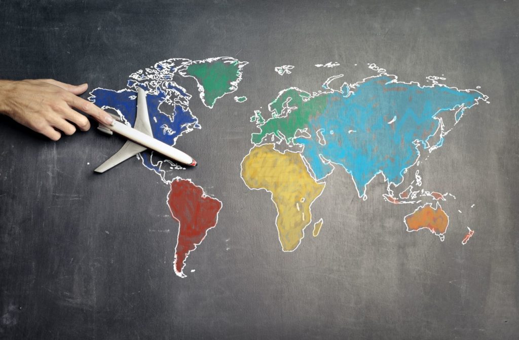 A grey background with a map of the countries drawn in multi-colored chalk, and a hand reaching out with a toy airplane. The featured image for Sky Bird Travel & Tours travel blog article "Misconceptions of Airline Consolidators."