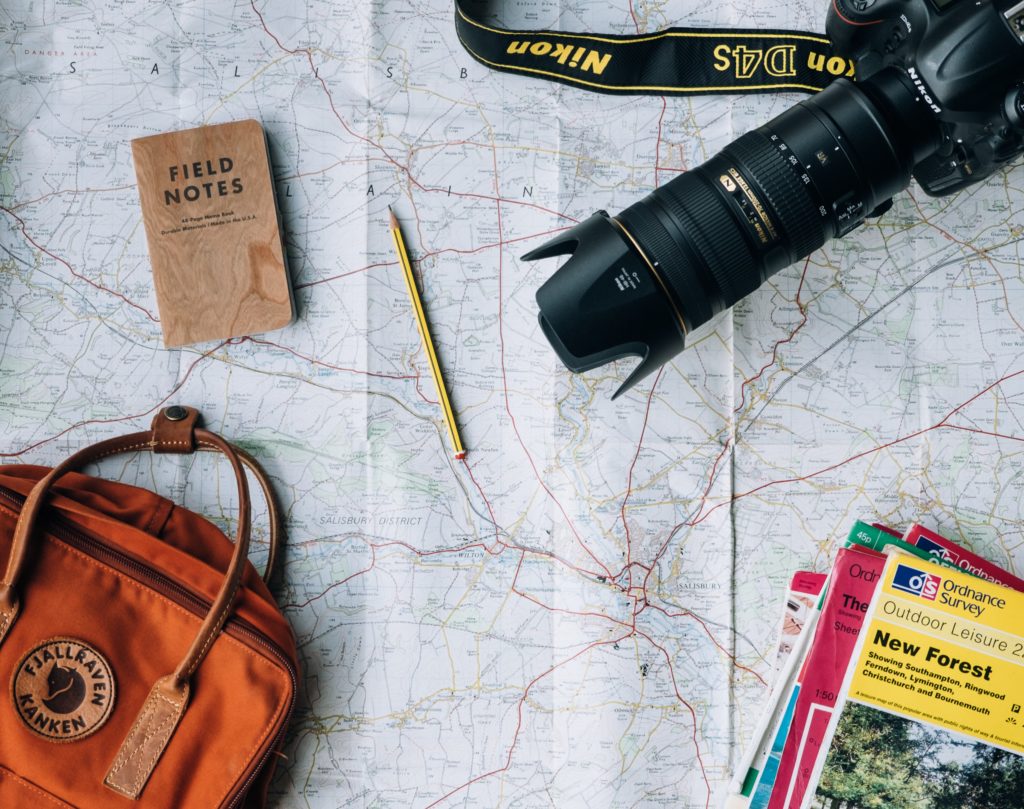 An image taken from above of a map laid out over a table, and a camera, travel articles, and a backpack lay on it. The featured image for Sky Bird Travel & Tours travel blog piece Month to Month Vacation Guide.