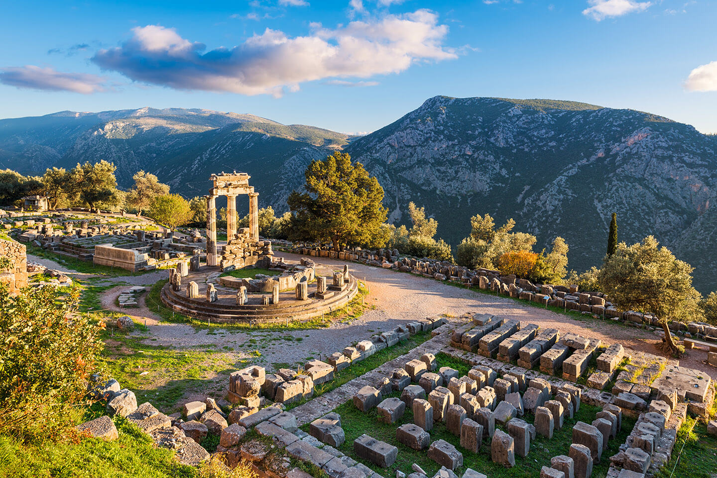 Ancient ruins and tall mountains on a sunny day in Delphi, Greece. There are the remains of great stone creations with light shining on them and trees everywhere.