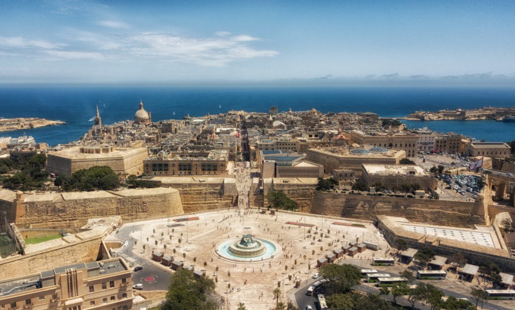 The town square of Valletta, Malta on the Mediterranean coast, with the blue ocean in the foreground. The square is a gathering place with a fountain and many people from Sky Bird Travel & Tours are surrounding it on their vacation.