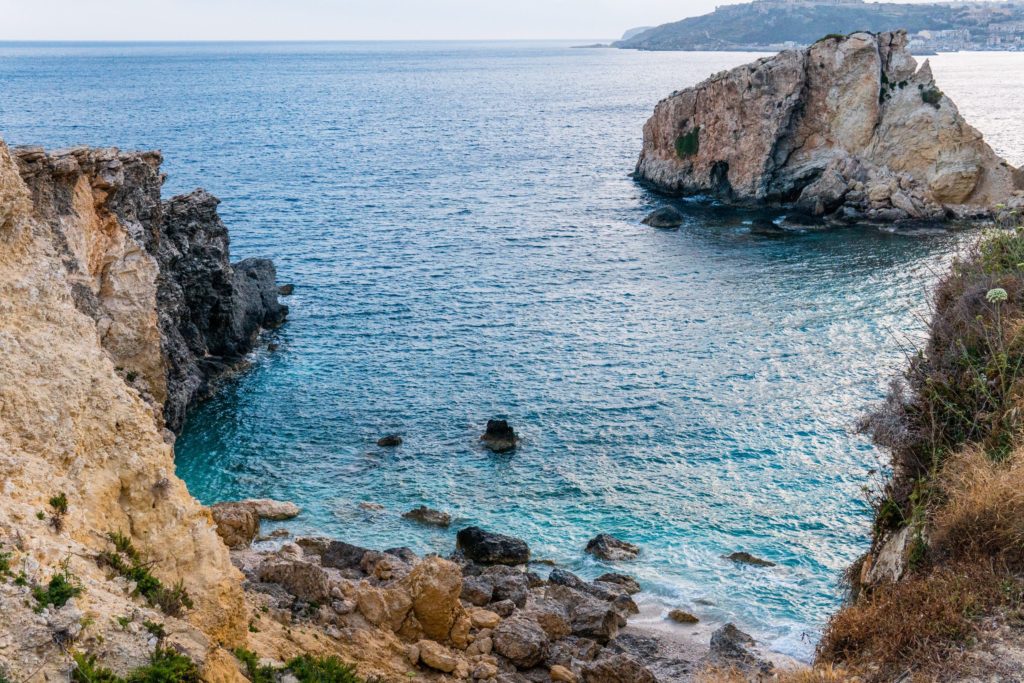 A travel photo of the blue ocean crashing onto the stone rock cliffs of Gozo Beach, on the island of Malta in Italy. Sky Bird Travel recommends traveling here, as it's a top 10 destination of 2023.
