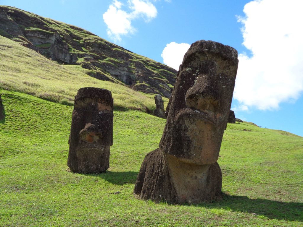 An image of Easter Island, with the bright blue sky and green hills behind two ancient mori head statues sitting in the ground. This can be seen on a customized tour from Sky Bird Travel & Tours.