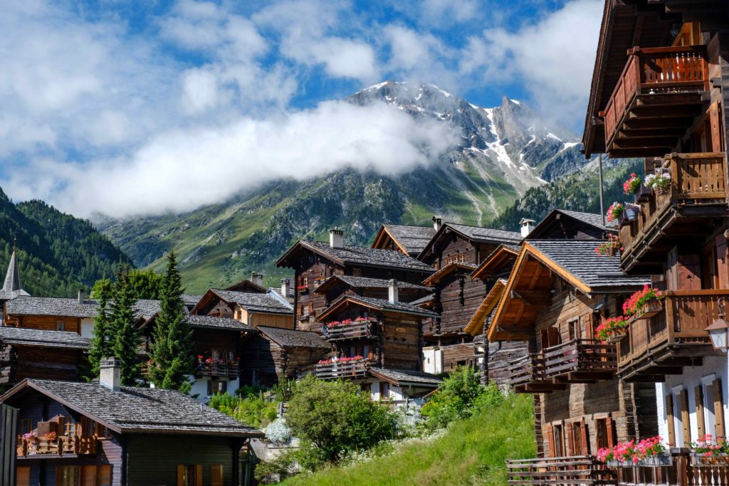 A beautiful picture of a small village in Switzerland and the Swiss Alps hidden behind clouds. The houses are wooden and traditional, and can be booked on sky bird travel & tours website.