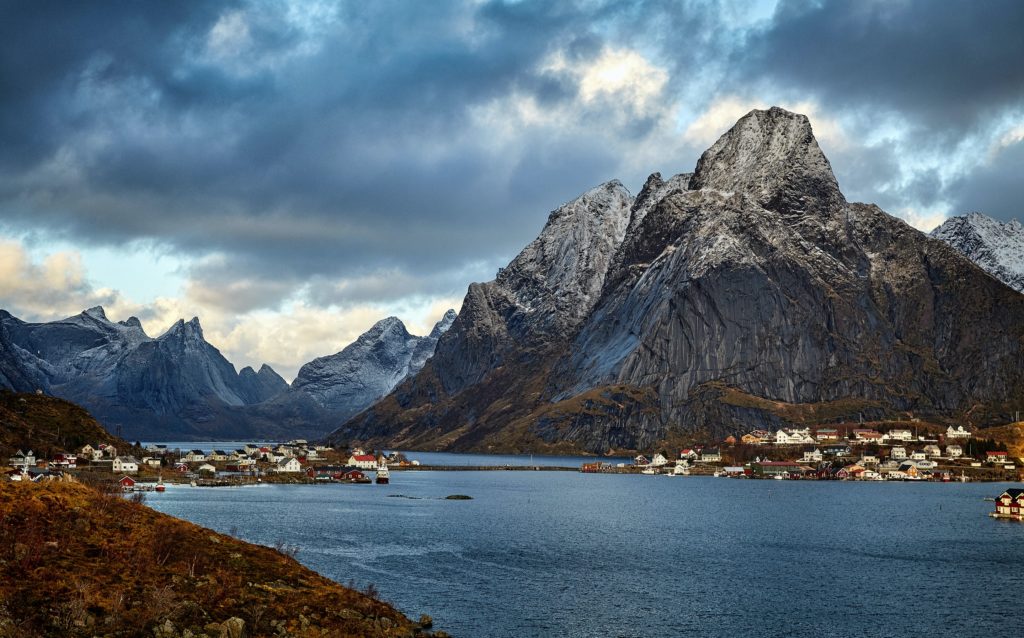 A wide landscape photograph of a gigantic mountain behind a winding river studded with white village houses. Sky Bird Travel & Tours promotes travel to Scandinavia in the article Month to Month Vacation Guide.