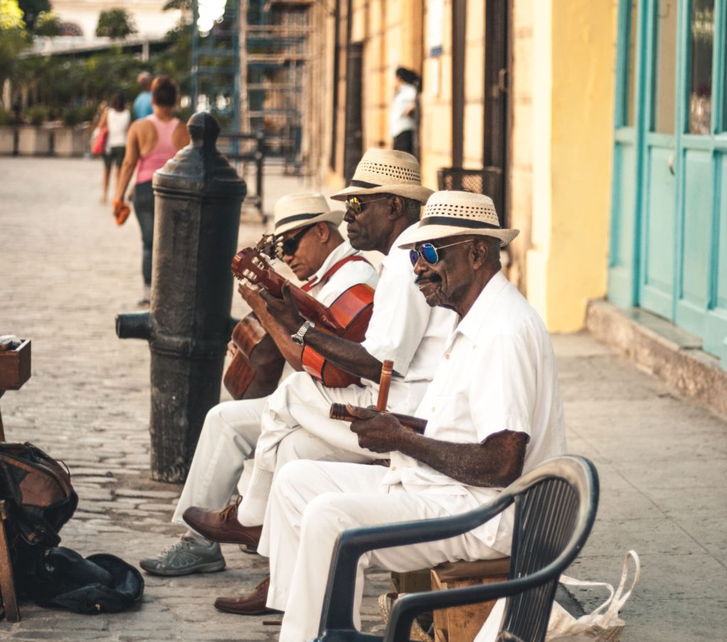 A candid image in Havana, Cuba of hreet men sitting on chairs and playing musical instruments to the other people in the street. The men wear matching white shirts, aviators, and fedoras.