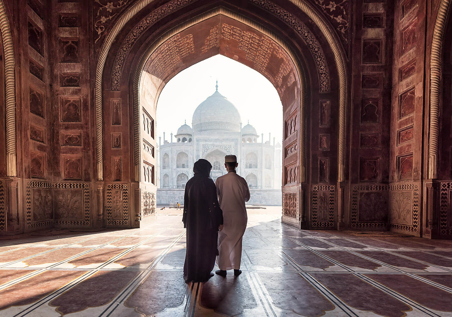 A young couple face away from the camera and pass under a large stone walkway to enter the stunning Taj Mahal in India.