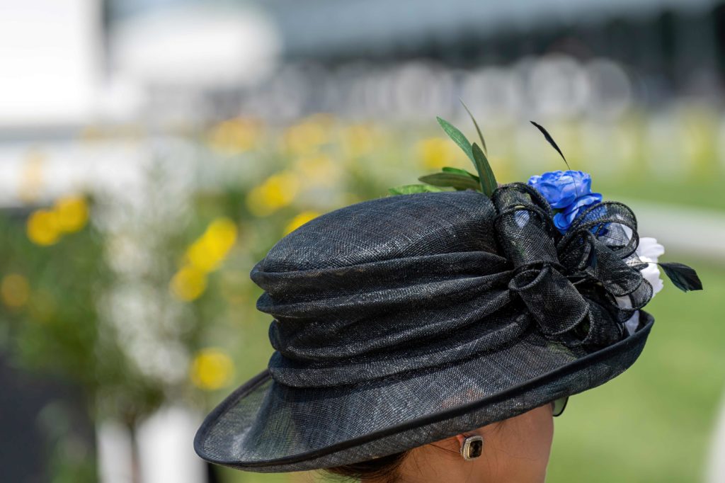 A photo taken from behind a woman at the Kentucky Derby in Louisville, and she is wearing a large and gaudy black hat with blue and white flowers. The travel agent was able to get a ticket to this top 10 spring break destination because of Sky Bird Travel & Tours.