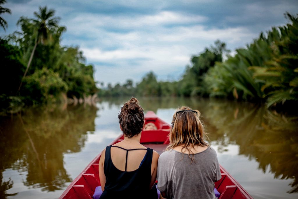 A travel photo of two young girls taken from behind in the Green Cathedral, in Kampot, Cambodia. The travelers are on a boat tour, booked with Sky Bird Travel & Tours, passing by green palm trees.