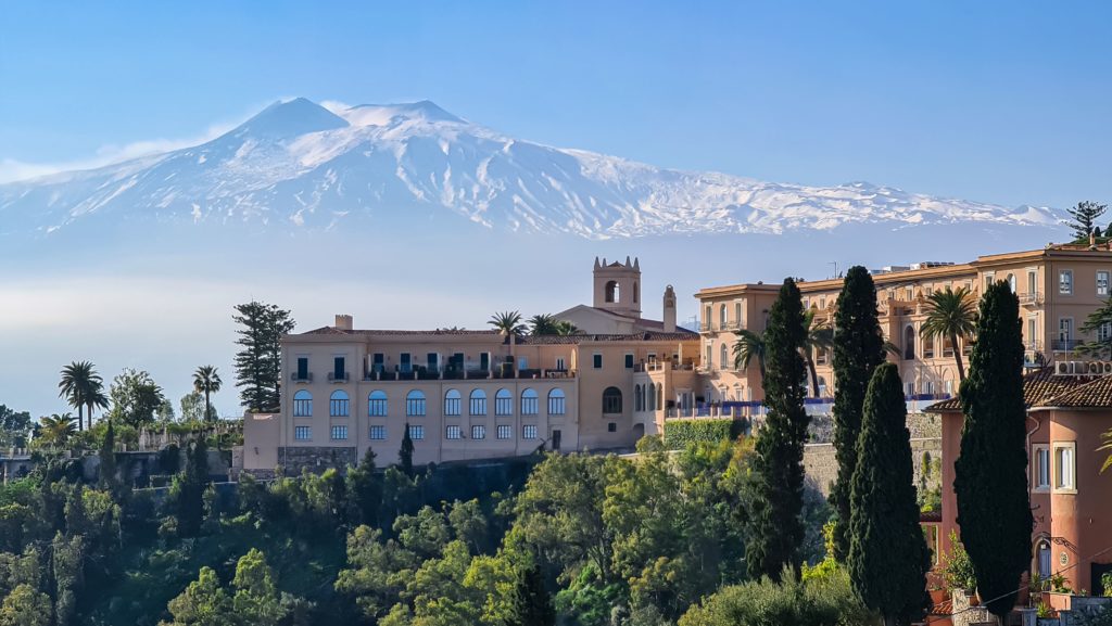 A beautiful landscape shot of the active volcano Mount Edna and the San Domenico Palace, which looks like an ancient Italian castle. This is the film location of The White Lotus season 2, which is a location that fans of the show can visit in real-life.