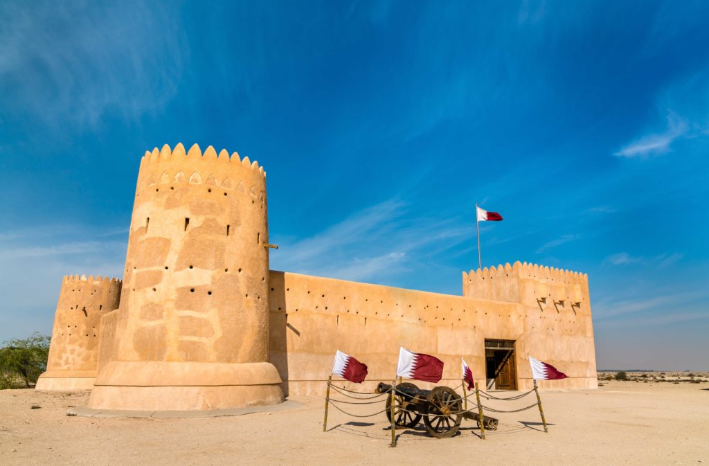 A large sandstone fort sits tall on the dirt floor while the bright blue sky lights up the image, and a small cannon is on display in front. This is the Al Zubara Fort in Doha, Qatar and is used in Sky Bird Travel & Tours blog Top Destinations Qatar for Clients.