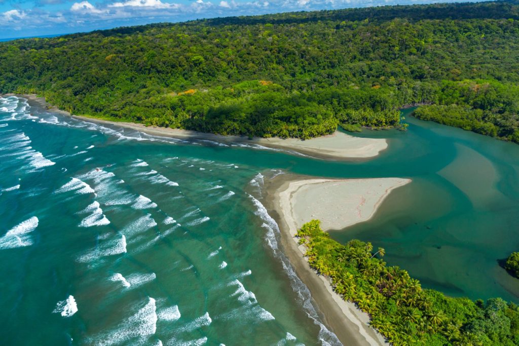 A stunning nature photograph of Corcovado National Park, in Puntarenas, Costa Rica. There is an island covered in bright green trees with the ocean crashing onto the small white beaches. In the Sky Bird Travel & Tours travel blog, this has declared this a Top 10 National Park to visit in 2023!