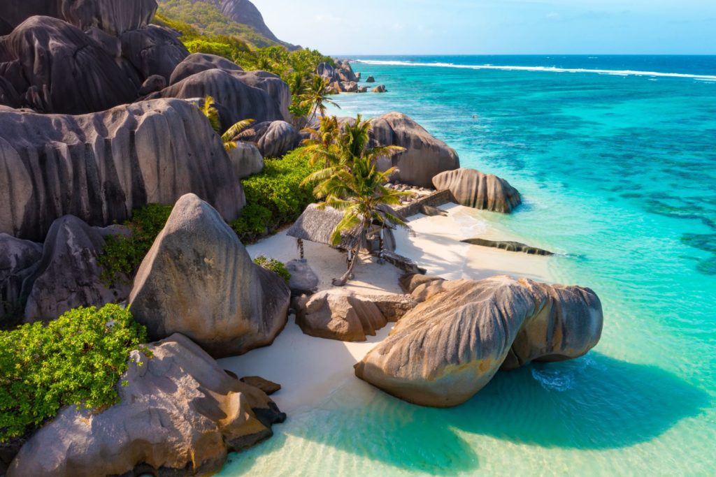 The beautiful beach, Anse Source d'Argent Digue, in Seychelles is the perfect vacation spot making it a top 10 destination of 2023. Large, smooth, grey boulders are scattered around a sandy beach, palm trees, and the stunning blue ocean.
