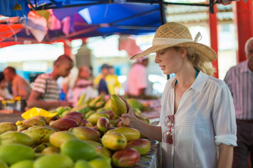 The fresh produce stalls of the infamous Victoria market in Seychelles, declared a top 10 destination for 2023 by Sky Bird. A woman wearing a straw hat stands at a produce stall, inspecting a fresh mango and the assorted fruits around her.