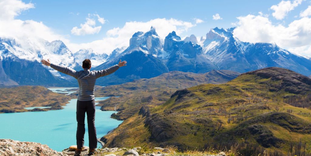 A young man stands with his back to the camera, facing a bright blue lake and the Patagonian Mountains with his arms outstretched. This legendary national park, Torres del Paine National Park in Puerto Natales in Chile.