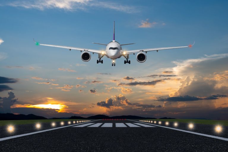 A large white plane is descending to the ground and is going to land on a runway, lit up by lights around the airport. This flight is carrying travel agent's clients to their various destinations, and earning travel agents amazing commission.