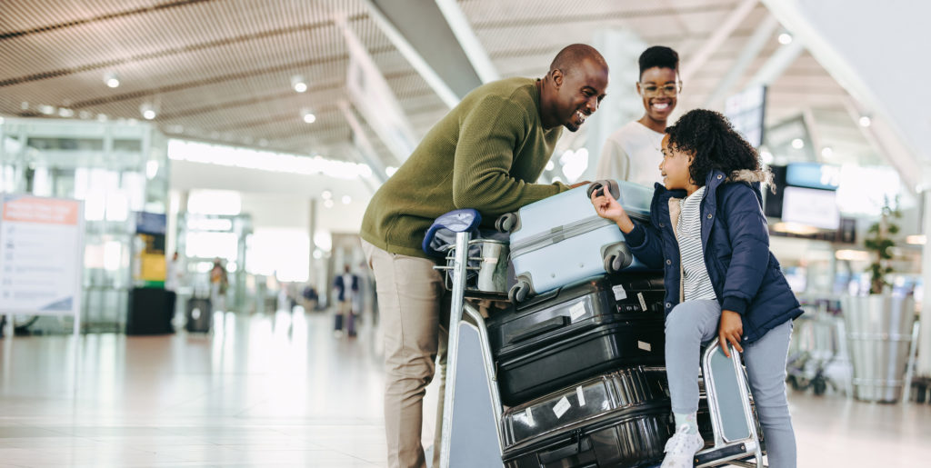 In an airport, a man and woman smile at their young daughter while pushing a luggage trolley to their departure or arrival gate. The featured image in Sky Bird Travel & Tours travel blog article for travel agents titled "Why You Should Always Book Client's Flights."