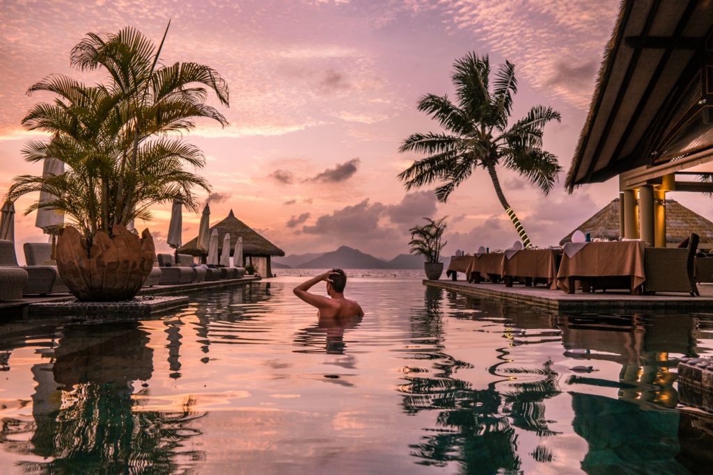 The featured image from Sky Bird Travel & Tours travel blog post, "Top 10 Destination: Seychelles." A young man is standing shoulder-deep in a hotel pool while watching a beautiful pink and purple sunset.