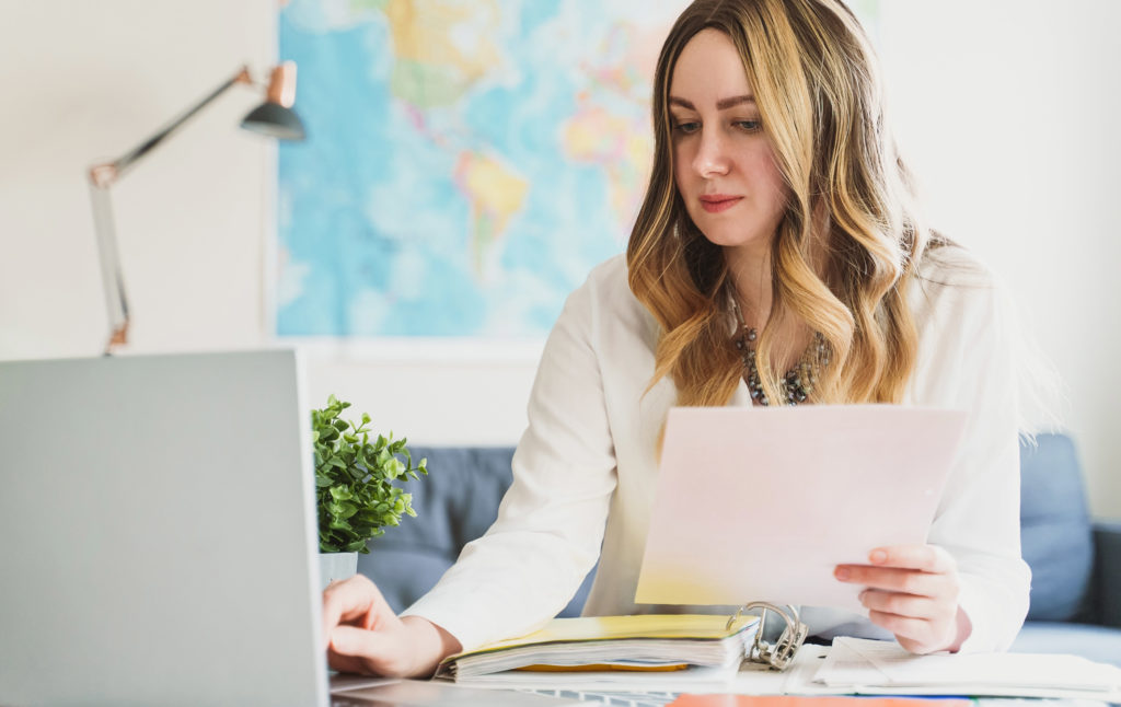 A young female travel agent looks down at the paper in her hand and binder on the table under her while using one hand to type on her silver laptop. This travel agent is booking airfare for their clients after reading Sky Bird Travel blog for the best tips for travel agents.