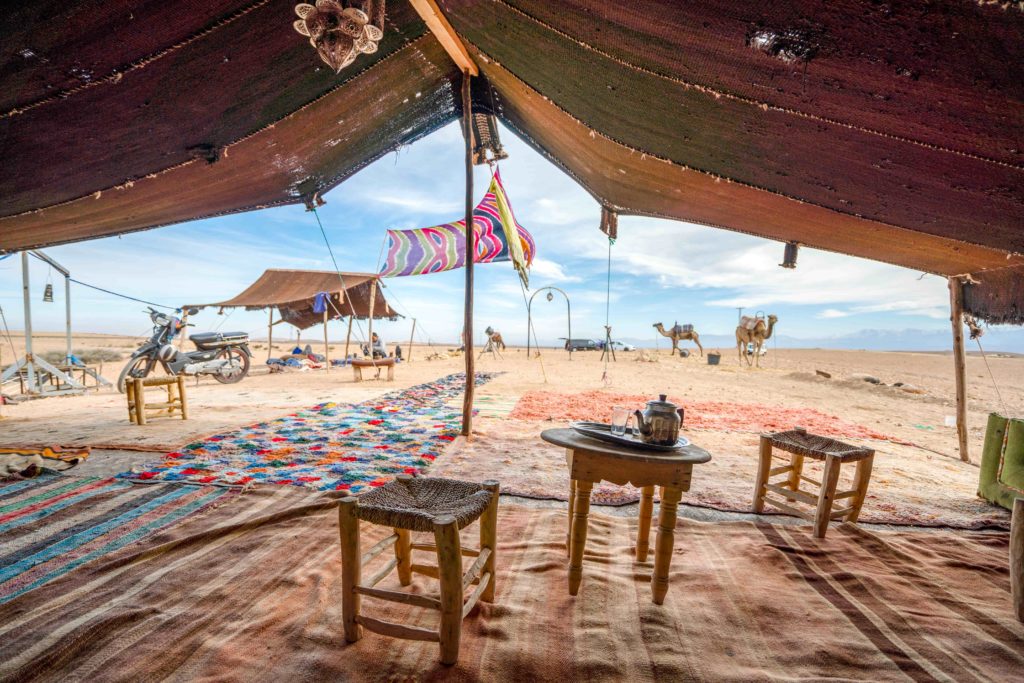 The view from inside a Bedouin tent in the Jordan desert looking out at camels and surrounded by middle east culture like tea and colorful carpets. This image is featured in the Sky Bird Travel & Tours tour guide blog article, "Top 10 Destination: Jordan," which lists the best things to do in Jordan!