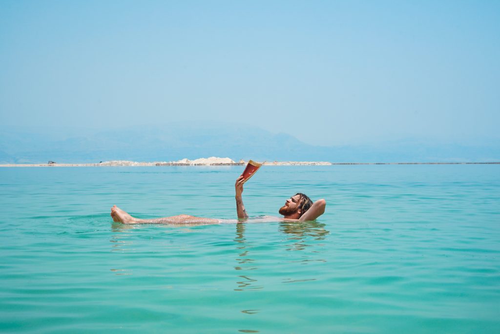 A calming shot of a man floating on the salty water in the Dead Sea and leisurely reading a book above the water. This image is featured in the Sky Bird Travel & Tours tour guide blog article, "Top 10 Destination: Jordan," which lists the best things to do in Jordan!