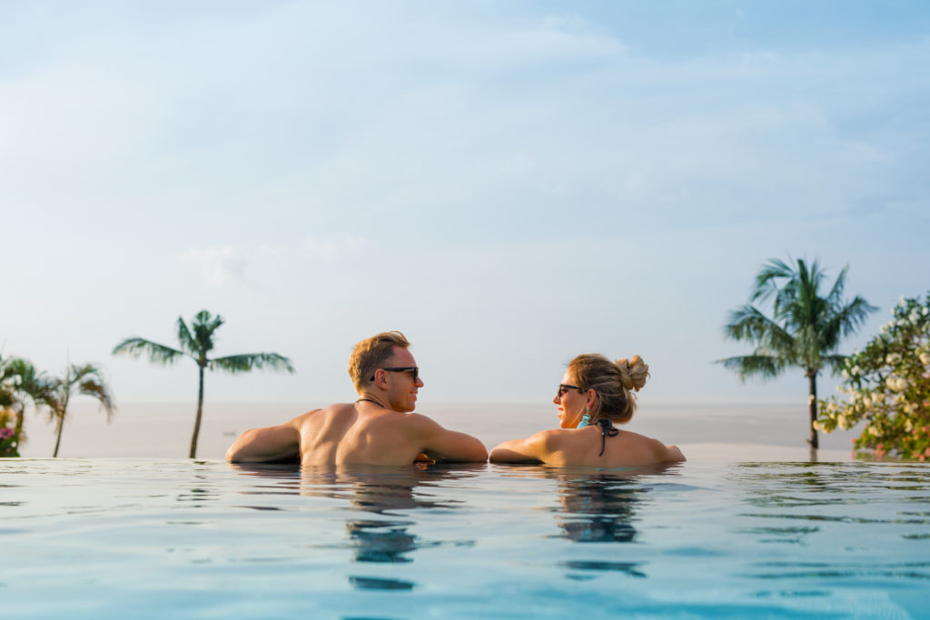 A man and a woman who just got married are on their honeymoon at a resort swimming in an infinity pool and falling in love. This is the featured image of the Sky Bird Travel & Tours blog, "Best Destinations for Honeymooners," which shows travel agents the most romantic locations for honeymoon!