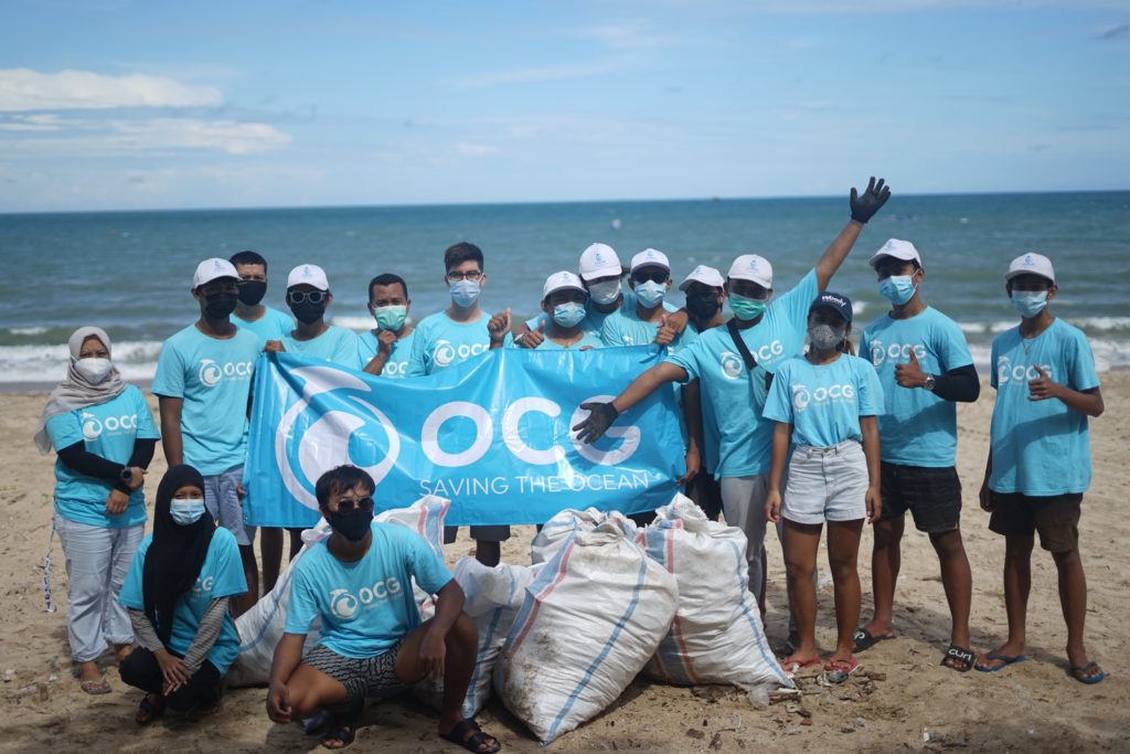 A large group of diverse volunteers pose on the beach with a bag of trash they cleaned up from the ocean and a banner that says saving the ocean. This image is featured in the Sky Bird Travel & Tours blog, "Encourage Safe and Responsible Travel," which shows travel agents sustainable travel practices!
