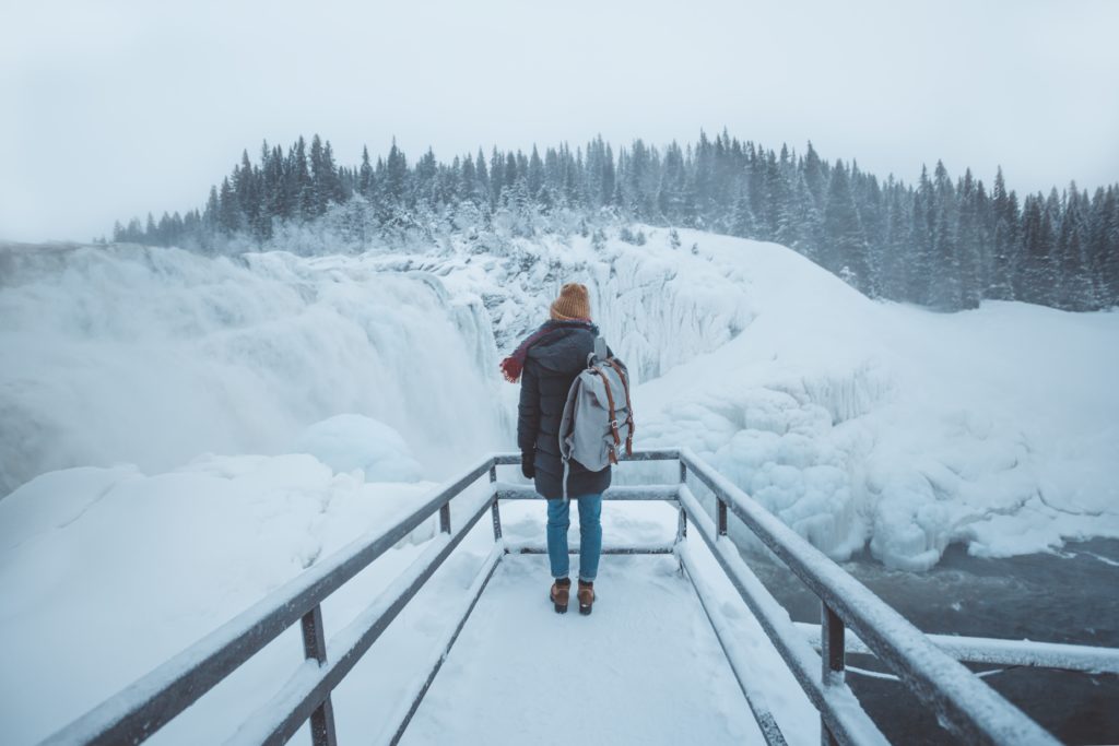 A young woman wearing a winter jacket, hat, and backpack stands facing a frozen waterfall and snowy forest in the winter. This image is featured in the Sky Bird Travel & Tours blog, "Encourage Safe and Responsible Travel," which shows travel agents how to guide their clients on a safe vacation!