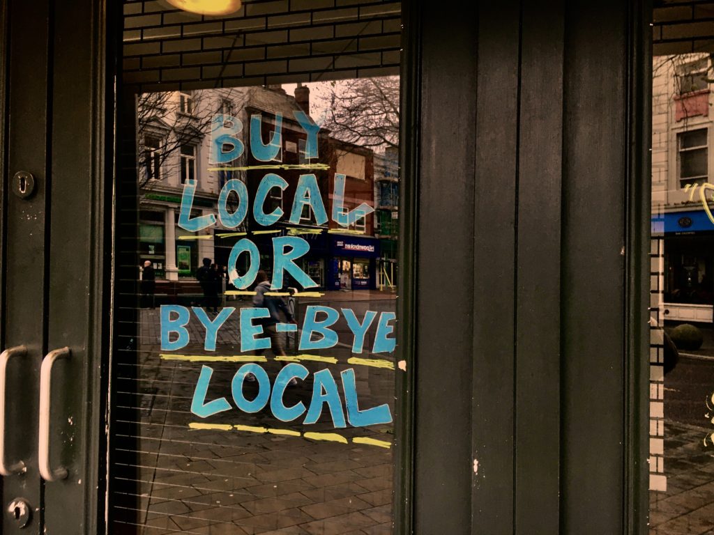 A close-up shot of a local store window with a hand-painted sign on the window saying "buy local or bye-bye local." This image is featured in the Sky Bird Travel & Tours blog, "Encourage Safe and Responsible Travel," which shows travel agents how to guide their clients on a safe vacation!