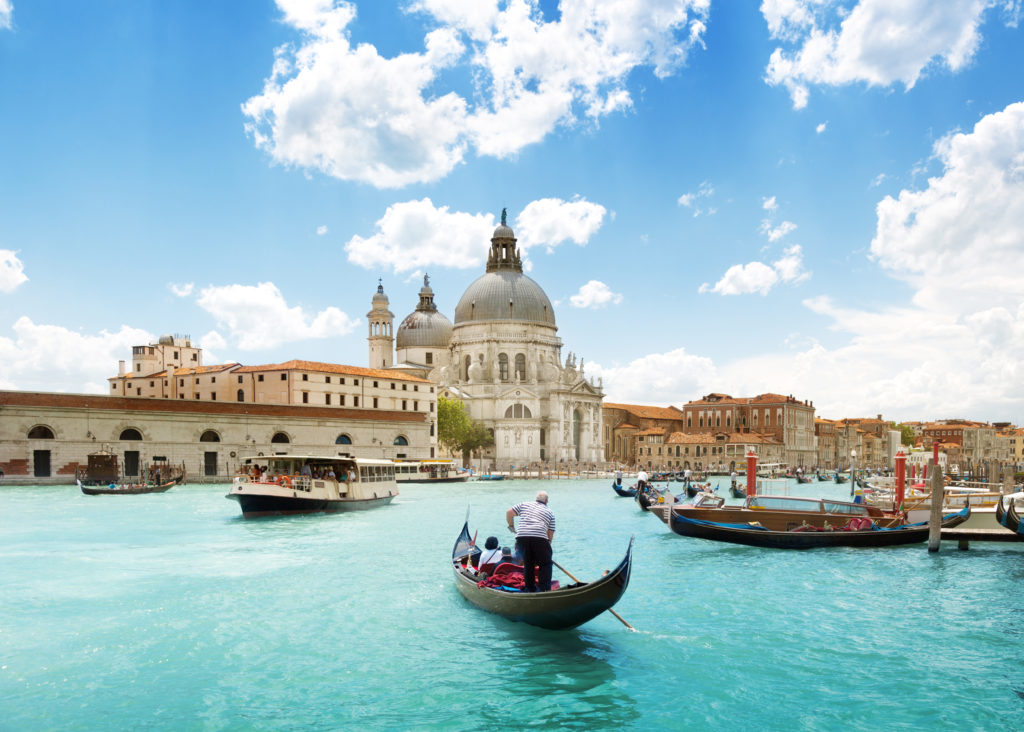 A beautiful image of the Grand canal and Basilica Santa Marina della Saltue with dozens of boats on the canals on a sunny day in Venice, Italy. This image is featured in the Sky Bird Travel & Tours blog, "5 Spots To Send Clients Before it's Too Late," which describes the top locations that are being permanently destroyed.