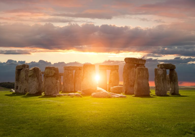A landscape of Stonehenge with the sun shining through the stone monument surrounded by a pink sunset. This image is featured in the Sky Bird Travel & Tours blog, "The Autumn Equinox," which describes the best places to view the fall equinox and how the festival started.