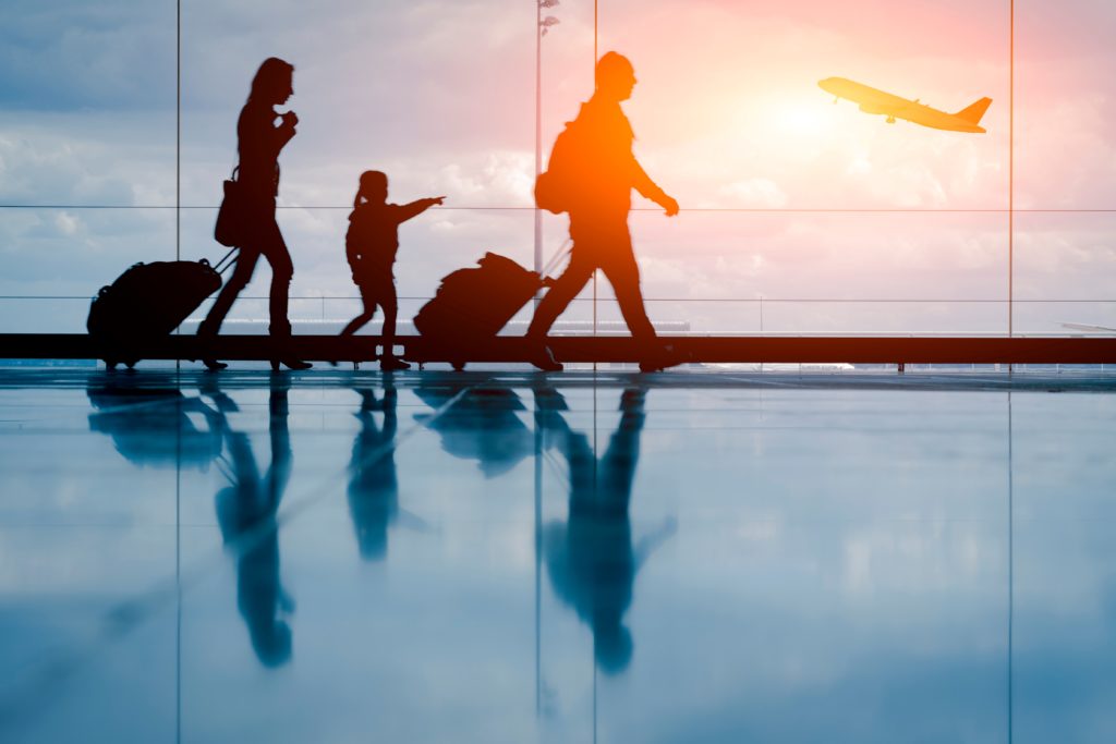 The silhouettes of a young family walking through the airport are highlighted by the sunset outside and a plane taking off. This image is featured in the Sky Bird Travel & Tours blog, "Booking Travel For Clients With Kids," which gives tips for booking family vacations with children.