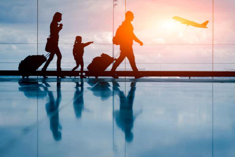The silhouettes of a young family walking through the airport are highlighted by the sunset outside and a plane taking off. This image is featured in the Sky Bird Travel & Tours blog, "Booking Travel For Clients With Kids," which gives tips for booking family vacations with children.