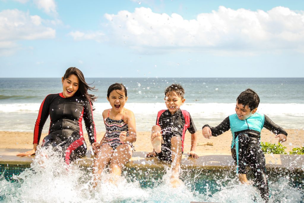 A mother, daughter, and two sons sit on the side of a pool on a tropical beach and kick their feet in the water. This image is featured in the Sky Bird Travel & Tours blog, "Booking Travel For Clients With Kids," which gives tips for booking family vacations with children.