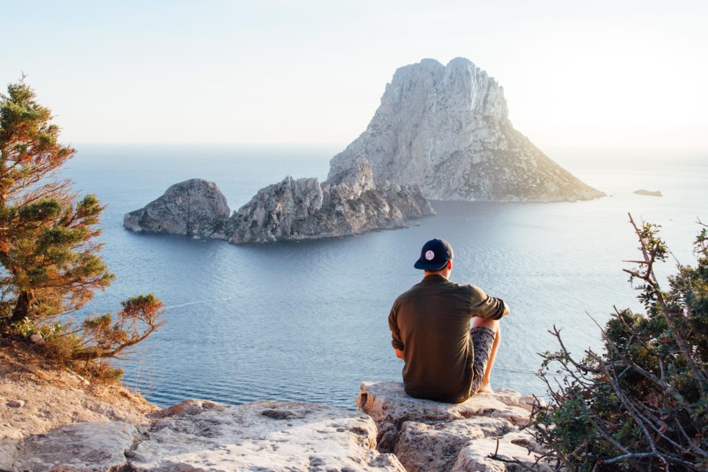 Featured in the blog "How To Celebrate World Tourism Day" by Sky Bird Travel & Tours, this image shows a man sitting on a cliff and looking at the sunset over the ocean.