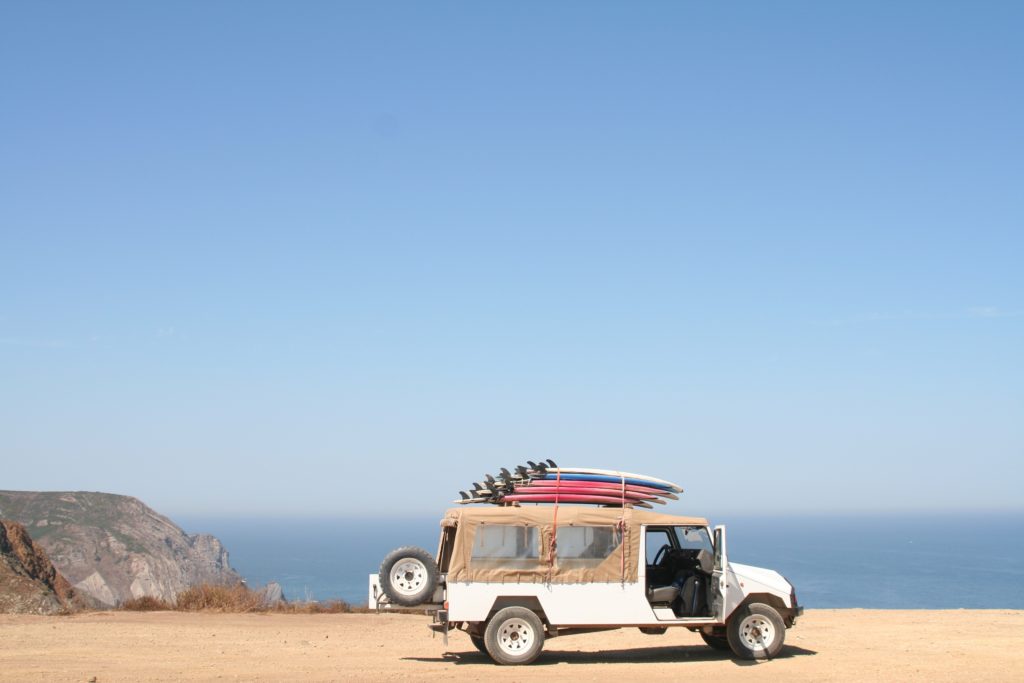 Featured in the blog "How To Celebrate World Tourism Day" by Sky Bird Travel & Tours, this image shows a van carrying a surfboard parked on the side of the road near a beach.