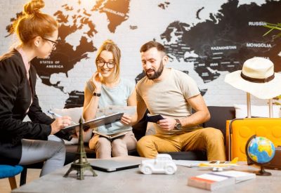 Three friends plan a trip, reviewing documents on a couch with a world map mural behind them and travel items nearby.