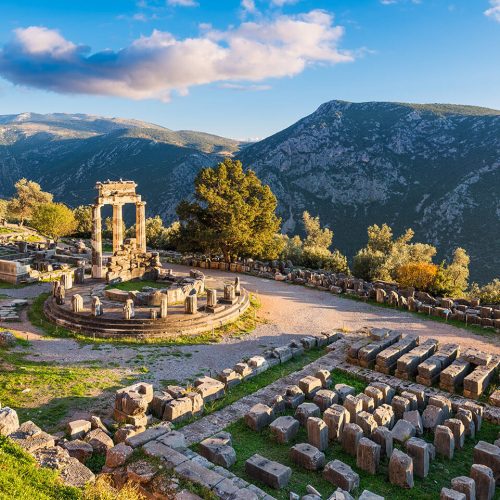 Ancient ruins and tall mountains on a sunny day in Delphi, Greece. There are the remains of great stone creations with light shining on them and trees everywhere.