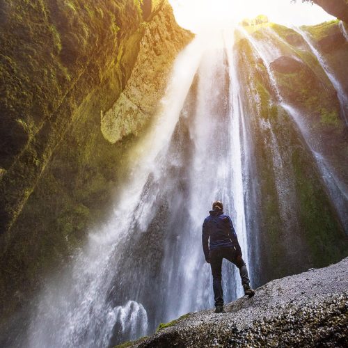 A student travel client in hiking clothes looks up at a large waterfall flowing inside of a cave on the side of a mountain in Iceland.